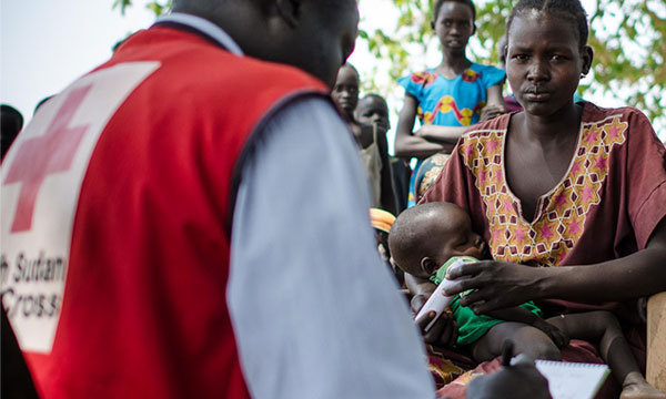 A Sudan Red Cross volunteer takes notes on a notepad, while a mother holds her baby.