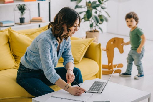 adult woman working on laptop with toddler playing nearby