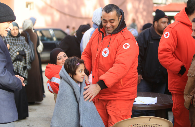 Dans une foule, un homme en uniforme de la Croix-Rouge pose une couverture sur les épaules d’une enfant.